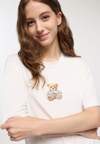 Biała, krótkosłębowa koszulka z haftowanym misiem na piersi, który nosi szary kokardę. Miękka tkanina o luźnym kroju.