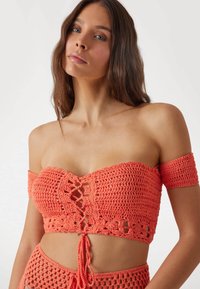 Koraal kleurige gehaakte crop top met off-shoulder mouwen, vetersluiting aan de voorkant en textuurpatronen. Licht en ademend materiaal, ideaal voor warm weer.