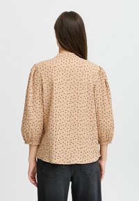 Blouse beige clair avec motif de cœurs noirs, dotée de manches ballon, d'un col froncé et d'un ourlet droit. Fabriquée en tissu léger.