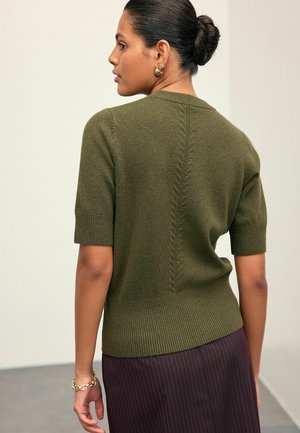 Vrouw met donker haar in een knot, gekleed in een olijfgroene gebreide trui met korte mouwen en een paarse streepjesskirt, staat voor een neutrale achtergrond.