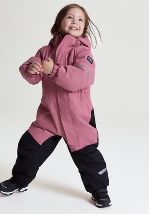 Unge barn smiler og bærer en pink og sort vandtæt flyverdragt med reflekterende detaljer, stående foran en ensartet lys baggrund.