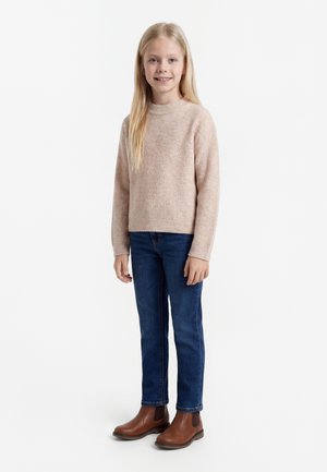 Ung jente med langt blondt hår iført beige strikkegenser, blå jeans og brune lærstøvler, står og smiler mot en hvit bakgrunn.