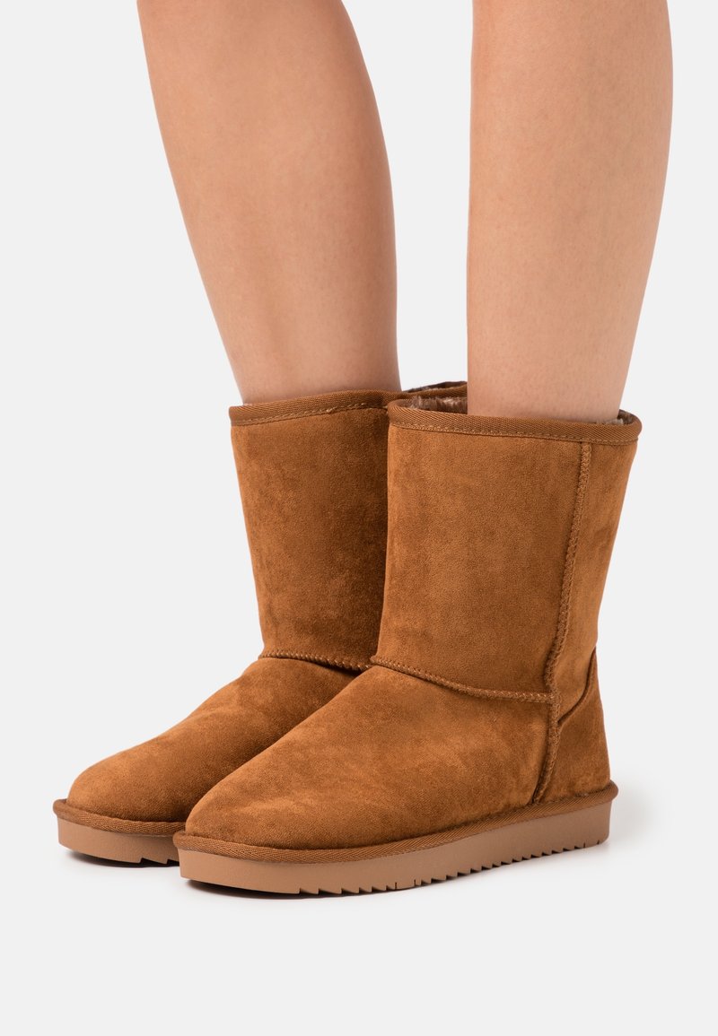 Des jambes portant des bottes d'hiver en daim marron mi-mollet avec des semelles en caoutchouc plates sur un fond clair uni.