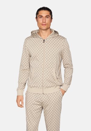 Mand iført beige hættejakke med lynlås og matchende bukser med sammenfletningsmønster, stående foran en ensfarvet baggrund.