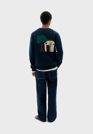 Navy sweatshirt featuring a decorative knitted design of a tree and house on the back. Paired with loose-fitting denim pants.