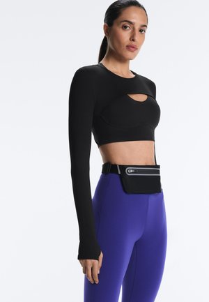 Kvinde iført sort langærmet cropped top med åbning ved brystet, lilla højttaljede leggings og sort taljeklips med lynlås.