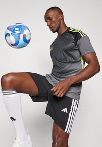 Maillot de sport Adidas en gris avec des accents noirs et verts fluo, associé à un short noir. Chaussettes et chaussures de soccer blanches. Ballon de soccer suspendu au-dessus.