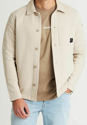 Homme portant une veste beige boutonnée par-dessus un t-shirt marron "CHASIN" accompagné d'un jean bleu clair, debout avec une main dans la poche.