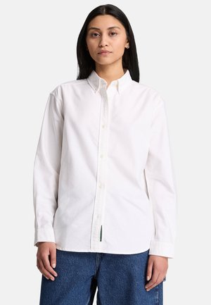 Kvinde med langt sort hår iført en hvid skjorte med knapper og blå jeans, der står mod en ensfarvet lys baggrund.