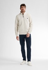 Jasno beżowa kurtka zapinana na zamek z fakturowanej tkaniny, wysoki kołnierz oraz czarnym logo na lewej piersi, w zestawieniu z ciemnoniebieskimi spodniami i białymi obuwiem.