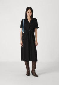 Vestido midi preto com mangas curtas, frente com botões, cinto na cintura e dois bolsos frontais. Combinado com botas castanhas até ao joelho e uma bolsa azul de ombro.