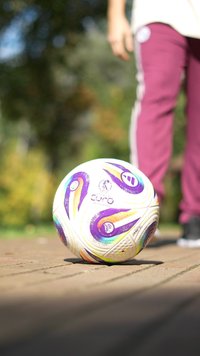 Futbolo kamuolys su baltu paviršiumi, puoštas spalvingais sūkurių raštais purpuro, oranžinės ir mėlynos spalvos; tekstūruoti paneliai ir aiškiai matoma prekinė ženklas.