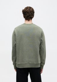 Mlad človek s kodrastimi lasmi, oblečen v olivno zeleno trenirko in črne hlače, stoji obrnjeno proti enostavni svetlo sivi ozadju.