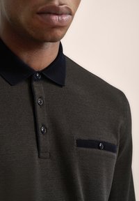Dunkelgrünes Poloshirt aus strukturiertem Stoff, mit navyfarbenem Kragen, drei Knöpfen und einer Brusttasche mit navyfarbenem Besatz und Knopfdetail.
