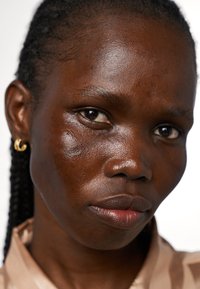 Gros plan d'un visage à la peau foncée, présentant une texture lisse, un éclat naturel, des cheveux tressés et de petites boucles d'oreilles en or.