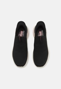 Fekete, fűző nélküli slip-on sportcipő, textúrált hálós felsőrésszel, világosszürke talppal és piros díszítéssel a betét logóján.