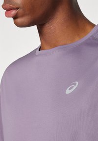 Lilla atletisk t-shirt med rund halsudskæring, korte ærmer og et diskret logo på brystet. Materialet virker glat og let.