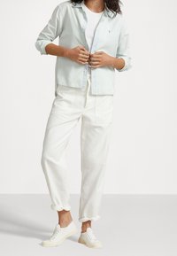 Ljusblå skjorta med knappar, vit linne, vita lösa byxor och vita sneakers med strukturerad sula, stående mot en enfärgad bakgrund.