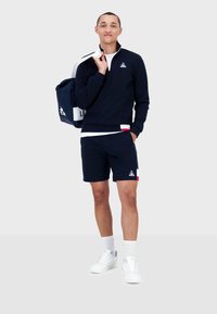 Felpa zip in navy con logo, pantaloni corti abbinati e sneaker bianche. L'outfit presenta un accento rosa in contrasto ed è abbinato a una borsa navy.