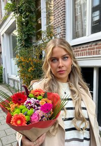 En bukett med blandade blommor i livliga färger, inklusive röd, rosa och grön, inlindad i tanfärgat och röd papper. Långa, smala blad framhäver arrangemanget.