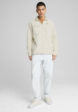 Lys beige lynlåsjakke med høj krave, skrå brystlomme og logo. Sammen med lyseblå løstsiddende jeans og hvide sko.