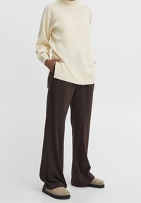 Pull en maille côtelée crème avec un col montant, associé à un pantalon ample marron foncé et des chaussures à enfiler couleur cognac. Design épuré et minimaliste.