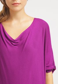 Lila, draperad topp gjord av mjukt tyg med kaskadkrage och korta, rullade ärmar. Slät textur med en avslappnad passform.