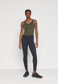 Ελαιόχρωμο αμάνικο μπλουζάκι με λευκές λεπτομέρειες, συνδυασμένο με μαύρα κολάν και μαύρα αθλητικά παπούτσια, στέκεται μπροστά σε απλό φόντο.