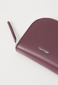 Bordowy skórzany portfel o gładkiej fakturze, zaokrąglonych krawędziach i wyraźnie widocznym srebrnym logo Calvin Klein z przodu.
