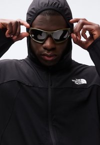 Άτομο με μαύρο μπουφάν με κουκούλα της North Face, φοράει και προσαρμόζει μεγάλα σκοτεινά γυαλιά ηλίου, με φόντο μια απλή λευκή επιφάνεια.