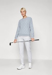 Pessoa com cabelo loiro curto a usar um suéter cinza, calças brancas e sapatilhas, segurando um taco de golfe horizontalmente à frente de um fundo simples.