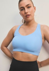 Vaaleansininen ribbed urheiluli bra, jossa on syvä kaula-aukko ja leveät hihnat, yhdistettynä korkeavyötäröisiin mustiin legginsseihin. Pehmeä, joustava kangas.
