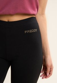 Leggings negros de tela elástica, con un logo "FREDDY" en pedrería dorada en la cadera izquierda. Textura suave y diseño ajustado.