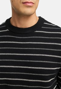 Sort strikket sweater med hvide vandrette striber, der har en ribbet rund halsudskæring og tekstureret stof. Detaljerne er klare og strukturerede.