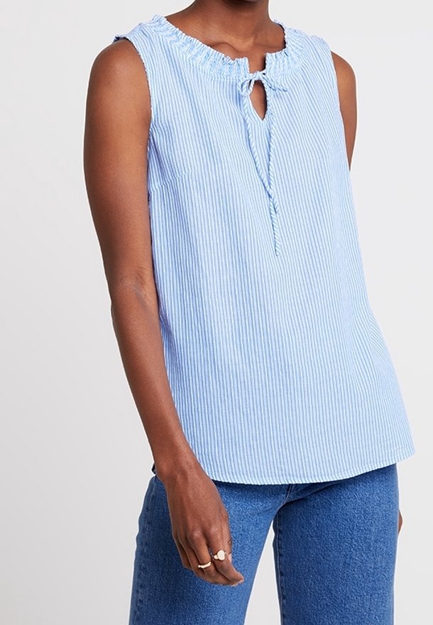 Top senza maniche azzurro chiaro e bianco a righe verticali con colletto arricciato e dettaglio con lacci, indossato con jeans blu da una persona.