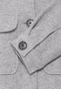 Sivý kabát z vlnenej zmesi s hladkou textúrou, vybavený dvoma veľkými tmavými gombíkmi a klopovým golierom; obsahuje predné vrecká s chlopňami.