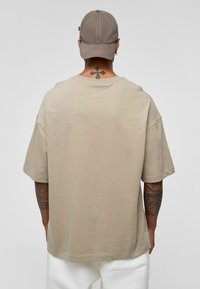 Beige oversized T-shirt met korte mouwen en een ronde hals, met een gladde textuur. Een grijze pet wordt gedragen en er is een tatoeage zichtbaar op de hals.