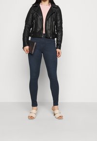 Chaqueta de moto de cuero negro, top rosa, Jeans ajustados azul oscuro y mules abiertos beige. La modelo sostiene una cartera oscura. Fondo simple.