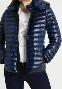 Navy puffer jacket with a glossy finish, featuring a zip closure, a detachable hood, and horizontal stitching. Paired with a printed white shirt.