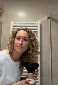 Woman with curly hair in white shirt holding black and gold facial steamer in a beige tiled bathroom with towel radiator behind her.