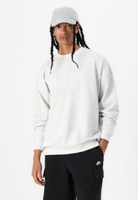 Grå sweatshirt med lange ærmer og ribbede manchetter, parret med sorte bukser med en cargo-lomme og broderet logo. Grå cap bæres oprejst.