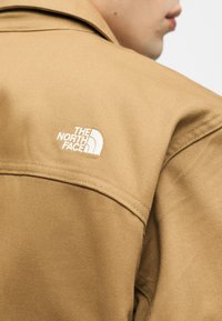 Κοντινό πλάνο ατόμου που φοράει μπεζ τζάκετ με λευκό κεντημένο λογότυπο The North Face στον ώμο, που δείχνει την υφή του υφάσματος και τις ραφές.