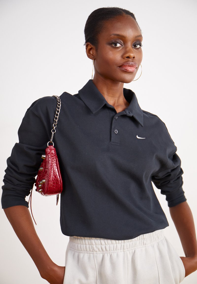 Camisa polo negra de manga larga con cuello y logo de Nike. Combinada con pantalones de chándal de color claro; modelo sosteniendo un pequeño bolso rojo texturizado.