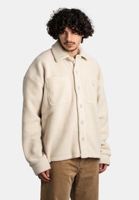 Beżowa polarowa kurtka typu shacket z wywijanym kołnierzem, dwiema kieszeniami na piersi, guziki z przodu i miękką teksturą. Nosi na sobie spodnie sztruksowe.