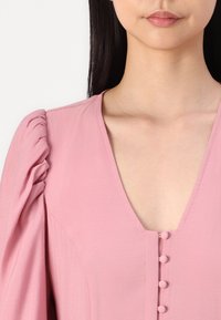 Vaaleanpunainen mekko, jossa on syvä V-kaula-aukko, puffihihat ja koristeelliset napit edessä. Valmistettu kevyestä kankaasta, jolla on sileä rakenne.