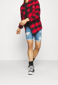 Camisa de franela a cuadros rojos y negros, camiseta negra, pantalones cortos de mezclilla azul desgastados y zapatillas negras con suelas y cordones blancos.