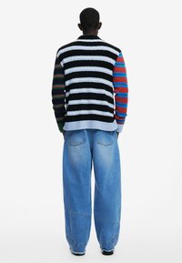 Stickad cardigan med svarta, vita och färgglada ränder; har mångfärgade ärmar. Kombinerad med löst sittande ljusblå denimbyxor.