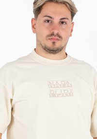 Beżowa koszulka z krótkim rękawem wykonana z miękkiej bawełny, z delikatnym, tonowym nadrukiem nazwy marki "NAPA PLIURI" z przodu.