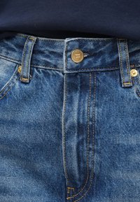 Μπλε τζιν παντελόνια με ψηλή μέση, με χρυσό κουμπί, κίτρινη ραφή και διακριτικές υφές. Κλασικός σχεδιασμός με πέντε τσέπες.