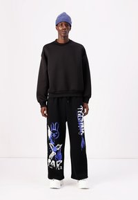 Svart genser med ribbestrikkede mansjetter og nederkant, kombinert med svarte vide bukser med blå grafiske design. Modellen har på seg en blå lue.